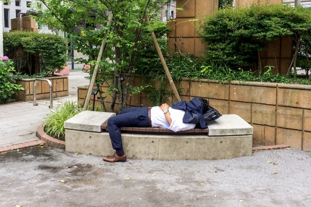公園のベンチで寝てサボっている、営業職の男性の写真